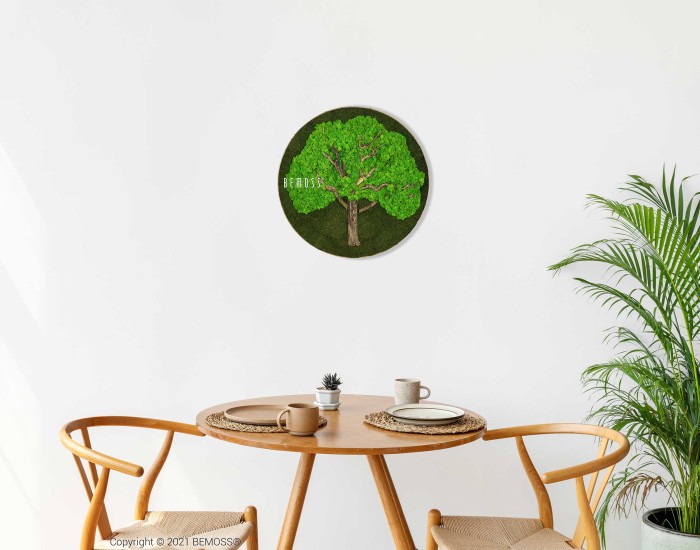 Een minimalistische eethoek is voorzien van een ronde houten tafel met twee stoelen. Op de tafel staan een kleine plant, een kaars en versieringen. Aan de witte muur hangt een Mosschilderijcirkel BOOM met een afbeelding van een boom met groen blad. In de hoek staat een potplant.