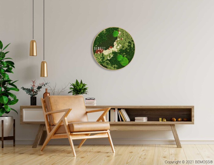 Een moderne woonkamer is voorzien van een houten fauteuil met lichtbruine kussens, een lage houten kast met open planken en een groene potplant. Boven de kast hangt een Mosschilderij cirkel BEMOSS® ORTHO ICE, wat een natuurlijk tintje geeft. Twee hanglampen dalen uit het plafond en werpen een warme gloed op het tafereel.