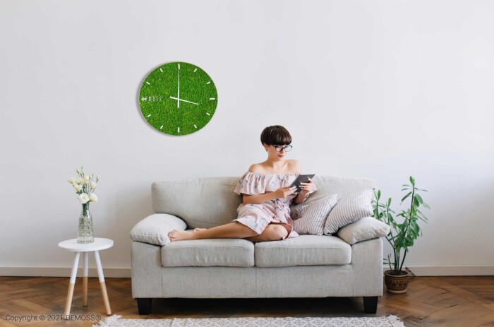 Een vrouw, gekleed in een zomerjurk, zit op een lichtgrijze bank en leest een tablet. De kamer heeft een witte muur met een Mos klok RENDIERMOS met wijzerplaat, een potplant en een kleine witte tafel met een vaas met bloemen. De setting is helder en minimalistisch.