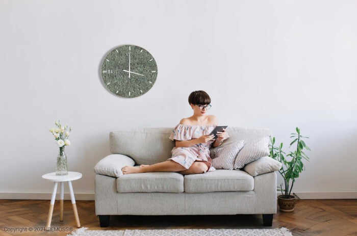 Een vrouw met een bril zit op een lichtgekleurde bank in een minimalistische woonkamer een boek te lezen. Aan de witte muur achter haar hangt een ronde groene Mos klok RENDIERMOS met dial. Aan de linkerkant van de bank staat een tafeltje met een vaas met bloemen en aan de rechterkant een potplant.