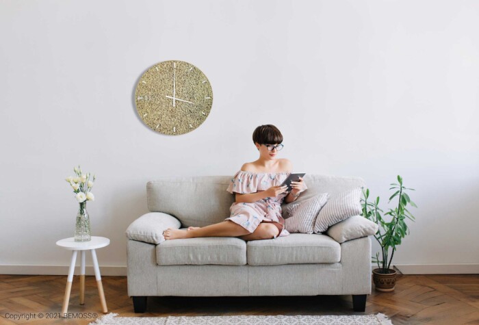 Een vrouw in een off-shoulder jurk zit op een grijze bank en kijkt naar een tablet. Ze heeft kort haar en een bril. De kamer is versierd met een klein wit bijzettafeltje met daarin een vaas met bloemen, een hoge vloerplant en een ronde wandklok met een gestructureerd ontwerp, naast een opvallende Mos klok RENDIERMOS met wijzerplaat aan de muur.
