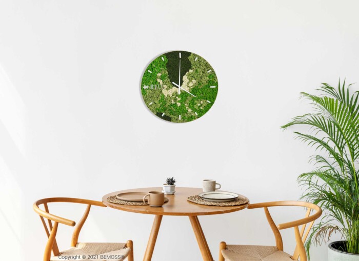 Een moderne wandklok met een groen, op de natuur geïnspireerd ontwerp dat lijkt op een Mos klok BEMOSS® ORTHO SPRING met wijzerplaat hangt boven een kleine ronde tafel. De tafel staat in een minimalistische kamer met lichtgekleurde muren. Er staan twee houten stoelen rond de tafel, waarop een kleine potplant en een mok staan. Rechts staat een hoge plant.