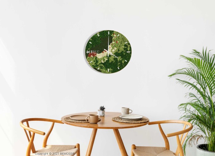 Aan een witte muur hangt een wandklok met een bloemmotief boven een klein, rond tafeltje en twee houten stoelen. Op de tafel staat een kleine potplant, een kopje en een kaars. Rechts van het tafereel staat een Mos klok BEMOSS® ORTHO SPRING met wijzerplaat, wat groen toevoegt aan de minimalistische setting.