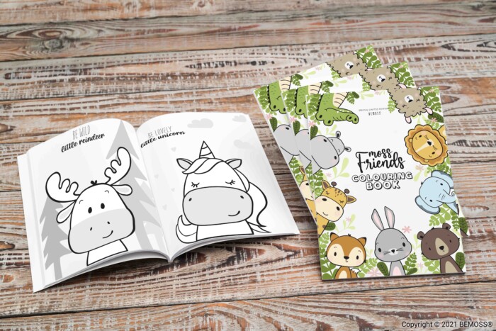 Een open kleurboek met cartoontekeningen van een rendier en eenhoorn ligt op een houten tafel naast een gesloten kleurboek met de titel 'Forest Friends Coloring Book', versierd met illustraties van verschillende bosdieren, evenals de serene groene tint van een BEMOSS-mosschilderij.