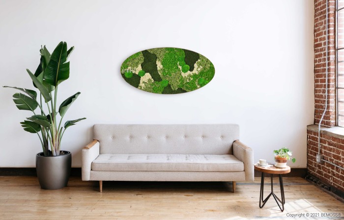 Een moderne, minimalistische woonkamer met een lichtgrijze bank, links een potplant en rechts een kleine houten tafel met een plant en een boek. Aan de witte muur boven de bank hangt een grote, ovale Mosschilderij Ellips BEMOSS® ORTHO TORRES. Aan de rechterkant bevindt zich een bakstenen muur met een raam.