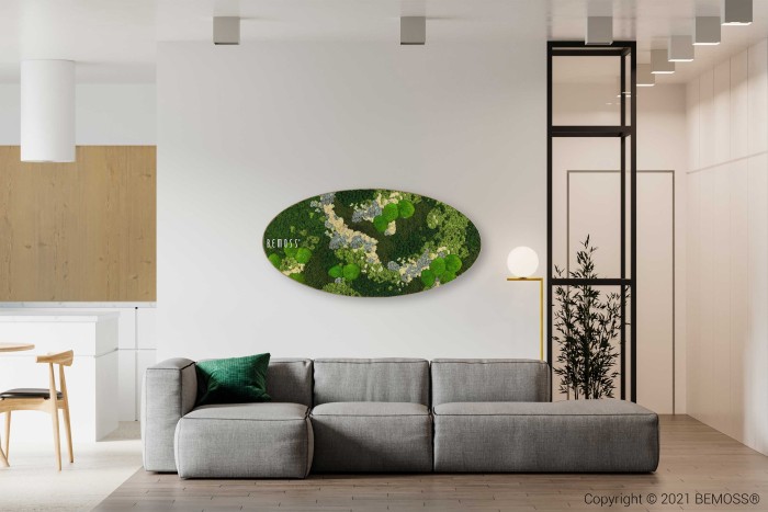 Een moderne woonkamer met een grijze sectionele bank, een groen kussen en een verticale wandlamp. Boven de bank bevindt zich een ovaalvormige Mosschilderij Ellips BEMOSS® ORTHO TORRES. De achtergrond bestaat uit een minimalistische keuken met witte kasten en een houten eettafel met stoelen.