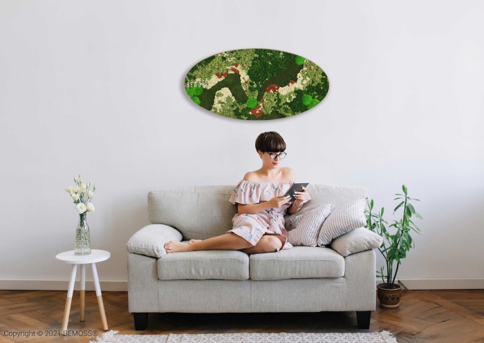 Een vrouw in een off-the-shoulder jurk zit op een lichtgrijze bank in een moderne woonkamer en leest een boek of tablet. Boven haar hangt een Mosschilderij Ellips BEMOSS® ORTHO TORRES met groene en rode abstracte ontwerpen. Potplanten en een tafeltje sieren de kamer en dragen bij aan de serene sfeer.