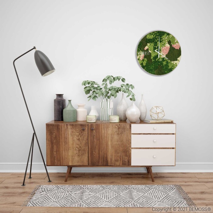 Een moderne woonruimte met een houten dressoir met witte laden, versierd met diverse decoratieve items, waaronder vazen, een plant en een geometrische sculptuur. Links hangt een strakke, zwarte vloerlamp, terwijl erboven een opvallende Mos klok BEMOSS® ORTHO SPRING in groene en roze tinten hangt.