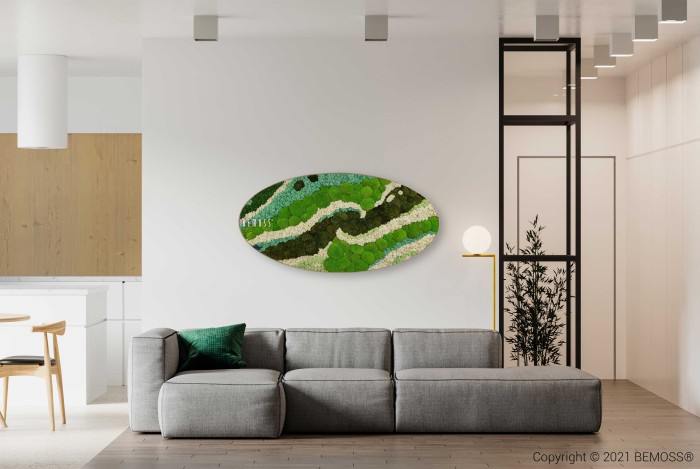 Een moderne woonkamer met een grijze hoekbank en een groen kussen. Boven de bank is een ovaalvormige Mosschilderij Ellips BEMOSS® ORTHO TORRES met groene en blauwe patronen op een witte muur gemonteerd. Naast de bank staat een minimalistische vloerlamp. Op de achtergrond is het keukengedeelte zichtbaar.