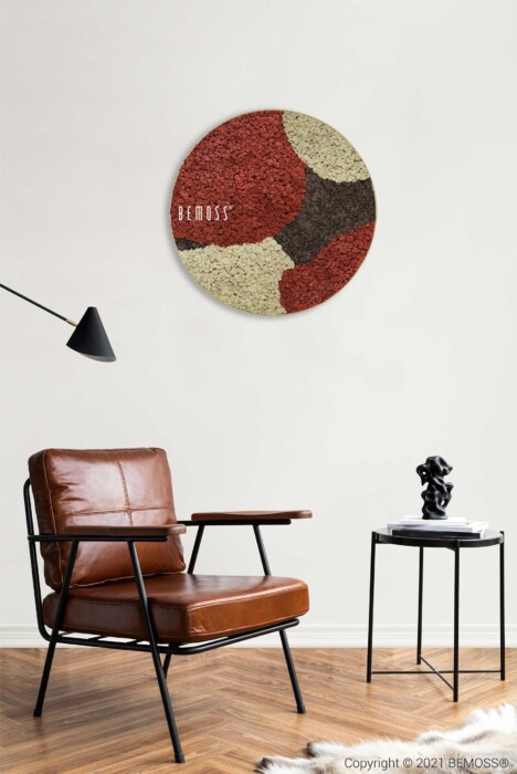 Een moderne woonkamer met een bruinleren fauteuil, een zwart bijzettafeltje met een beeldhouwwerk en een rond, abstract kunstwerk aan de muur met rode, beige en donkerbruine kleuren. Grenzend aan de kunst bevindt zich een Mosschilderij-cirkel BEMOSS® ORTHO SIENNA die een natuurlijke textuur toevoegt. Aan de linkerkant is een zwarte, schuine vloerlamp geplaatst. De vloer is van licht hout.