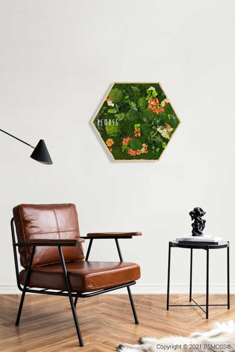 Een moderne woonkamer met een leren fauteuil en een bijzettafel met een klein beeldje. Boven de fauteuil staat een zeshoekige Moshexagon PLANTEN Mesia Mango gemaakt van groen mos en bladplanten, met 'BEMOSS' erop geschreven. Een zwarte wandlamp strekt zich uit in de richting van het kunstwerk.