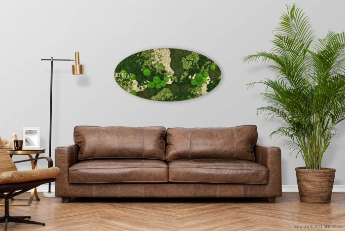 Een stijlvolle woonkamer is voorzien van een bruinleren bank, een potplant en een abstract ovaalvormig groen muurkunstwerk boven de bank. Links maken een gouden vloerlamp en een bijzettafel met fotolijsten de gezellige opstelling compleet. Een Mosschilderij Ellips BEMOSS® ORTHO OLVERA voegt textuur toe aan het subtiele chevronpatroon van de houten vloer.