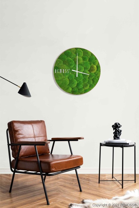 Een minimalistische woonruimte is voorzien van een groene Mos klok BOLMOSS Minerva met wijzerplaat op een witte muur, een bruinleren stoel met een zwart metalen frame en een klein zwart bijzettafeltje met daarop een modern beeldhouwwerk. Een zwarte wandlamp voegt een vleugje elegantie toe. De hardhouten vloer is gedeeltelijk bedekt met een wit tapijt.