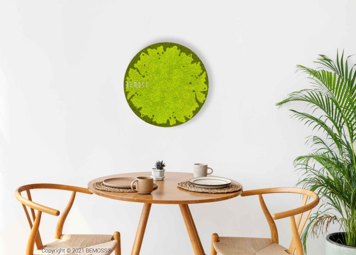 Een minimalistische eethoek is voorzien van een ronde houten tafel met twee bijpassende stoelen. Op tafel staan een mok, een theepot en borden op placemats. Aan de witte muur boven de tafel hangt een cirkelvormige Mosschilderij-cirkel BEMOSS® MARE, wat een elegant vleugje groen toevoegt. Rechts staat een potplant.