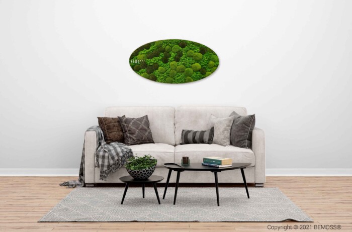 Een moderne woonkamer met een witte muur, met een grijze bank met diverse kussens. Een ronde, natuur-thema wandkunst hangt boven de bank. Voor de bank staat een ronde salontafel versierd met een kleine plant en spullen. De vloer heeft een lichtgrijs kleed.